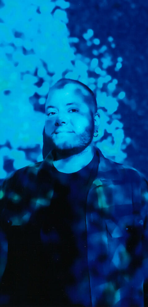 Elia is covered by a blue light and looks towards the camera. He wears a black t-shirt and a squared shirt.